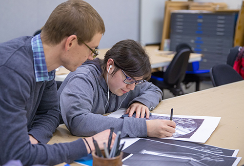 Art instructor looks on as an art student sits at a table working on a drawing