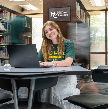 sa female student sits in a library with a laptop on a table with a blur behind her as if life is moving past her while she studies
